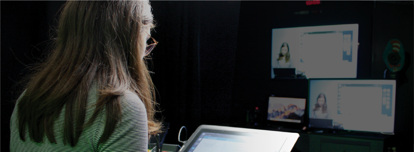 An instructor looks down at a digital control panel at a recording studio. Two monitors in the distance show a video feed of the instructor talking over slides.